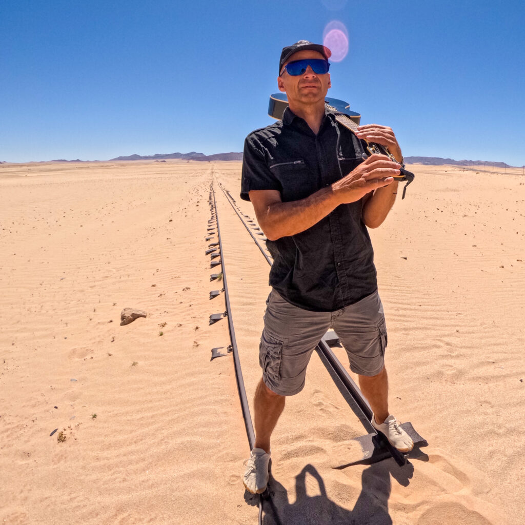 Person standing in the desert at the end of an old railway line with guitar. With writing: Expectations Cedric Vandenschrik